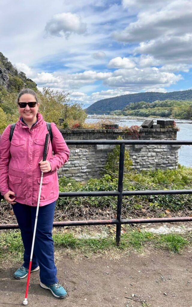 Vivian is standing in front of an overlook with the river, mountains, and clouds behind her. She is smiling and wearing a pink jacket, sunglasses, and holding her cane. 