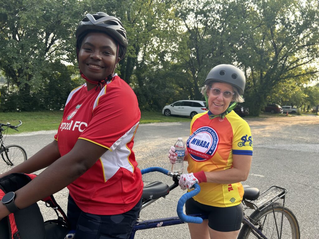 Mary and tandem captain Thembi stand with a tandem bike in Georgetown. Mary is wearing her MWABA bike jersey and smiling.