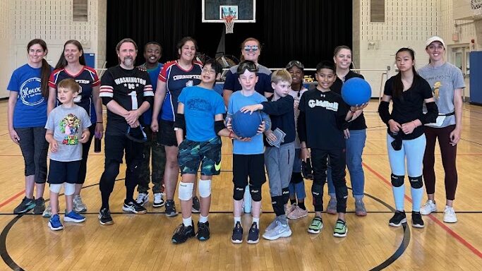 Coaches and youth participants pose with goalballs and in their pads at a youth goalball practice in May. From left to right are Coach Maureen, Coach Amanda, JP, Coach Kurt, Rahiem, Coach Lori, Eli, Calvin, Coach Hailey, William, Layla, Luis, referee Kristen, Cecily, and Coach Claire.