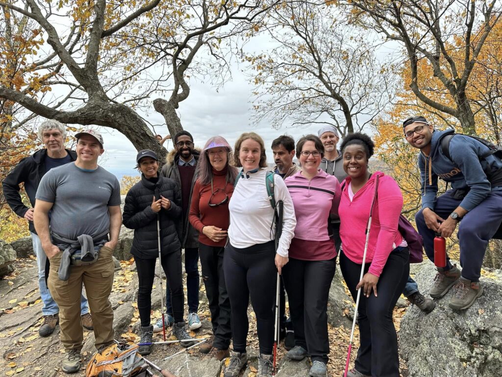 Karla (center, in a white shirt), stands with a group who went hiking at Catoctin in October 2022. The group stands poses at the lookout at Chimney Rock, flanked by trees with orange leaves. Starting with the third person from the left, the hikers and guides include Qudsiya, Sean, Carol, Karla, Marcos, Shira, Brian, Audrey, and Nafij.